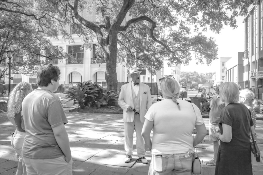 Group of people listening to a guide in a hat under a tree in a city plaza
