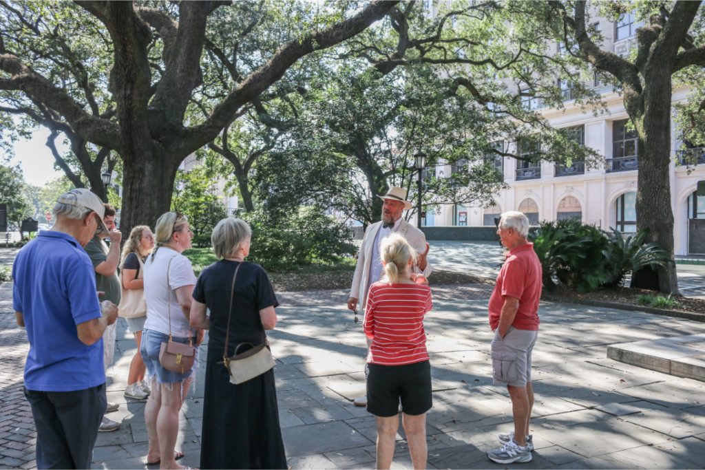 Group of tourists on a guided tour in a park, engaged with a guide under large trees near historical building