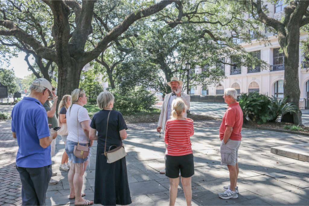 Tour guide leading a group of tourists through a park with large trees and historic building in the background