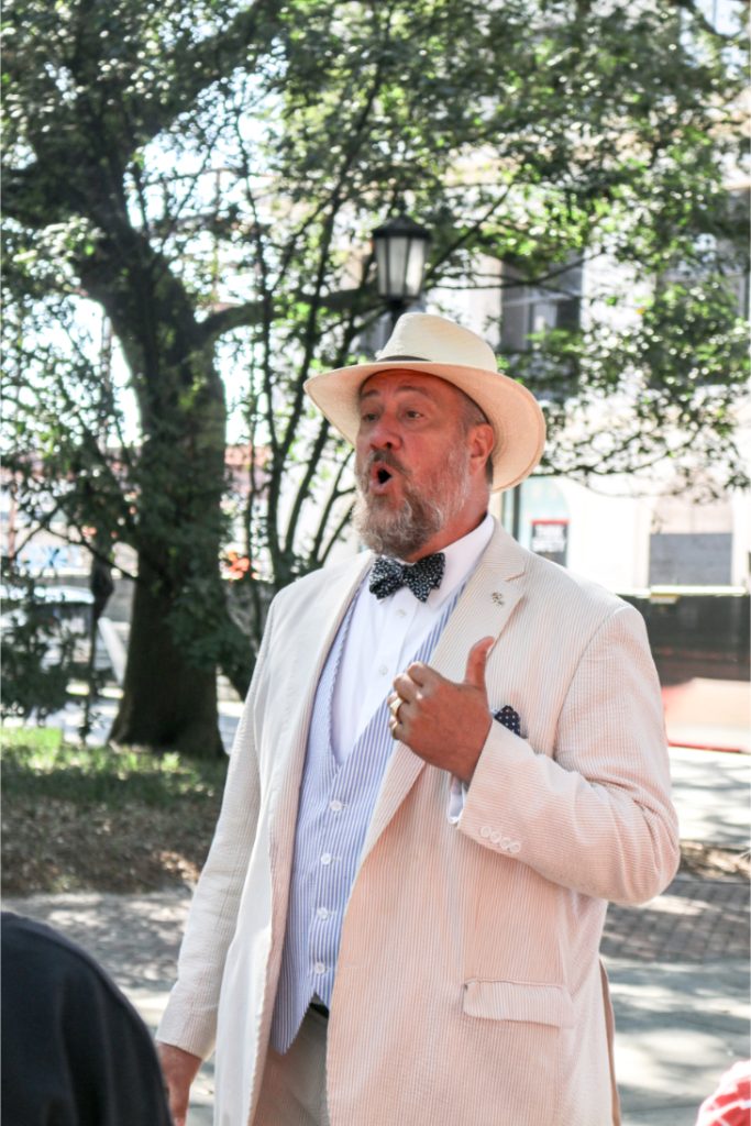 Man in straw hat and beige suit speaks animatedly outdoors under trees