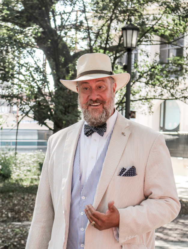 Bearded man in a stylish hat and suit with a bow tie standing outdoors on a sunny day near trees and historic buildings