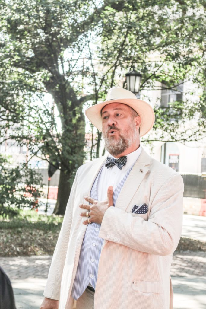 Man in vintage suit and hat gestures while speaking outdoors under trees