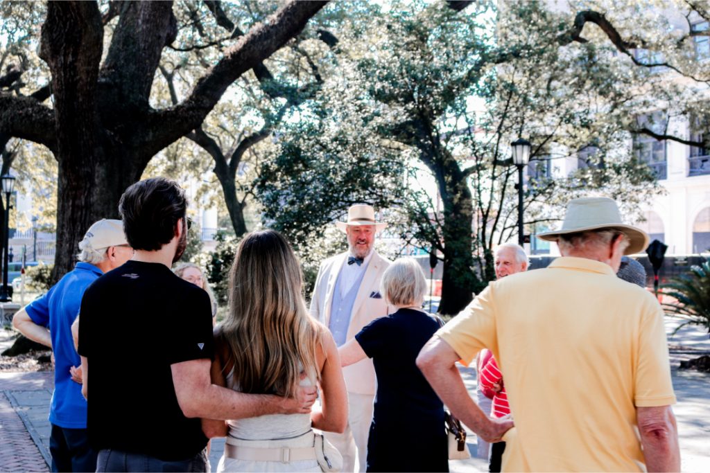 Group of people enjoying a sunny day in the park, with a man in a white suit speaking to them