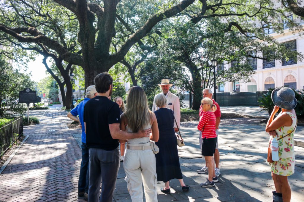Tour group exploring a historic park under large oak trees, listening to a guide in a sunny outdoor setting