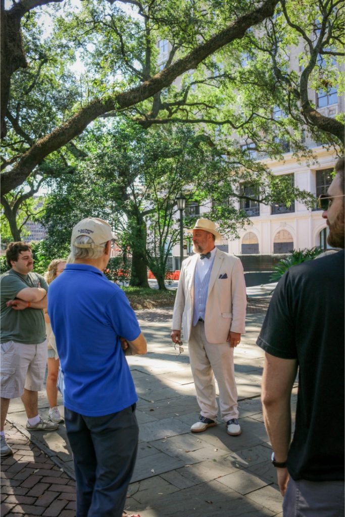 Tour guide in white suit leads a group discussion under shady trees in an urban park setting