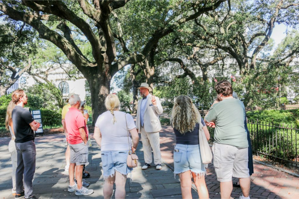 Tour guide in hat speaking to group under large trees in park setting
