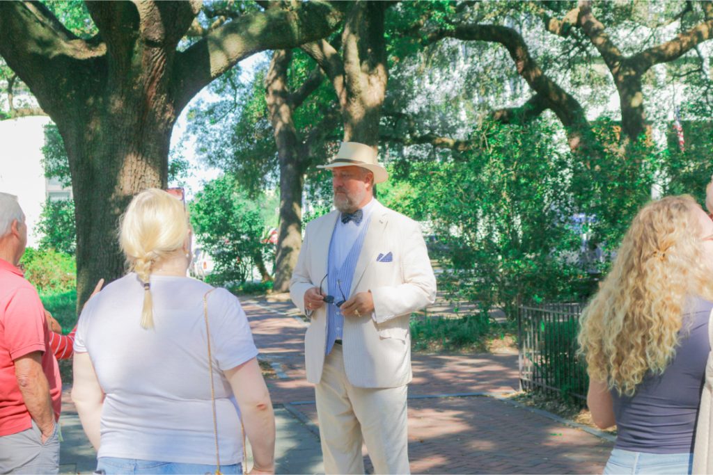 Tour guide in a suit and hat leads a group through a lush, tree-lined park walkway, engaging in lively discussion