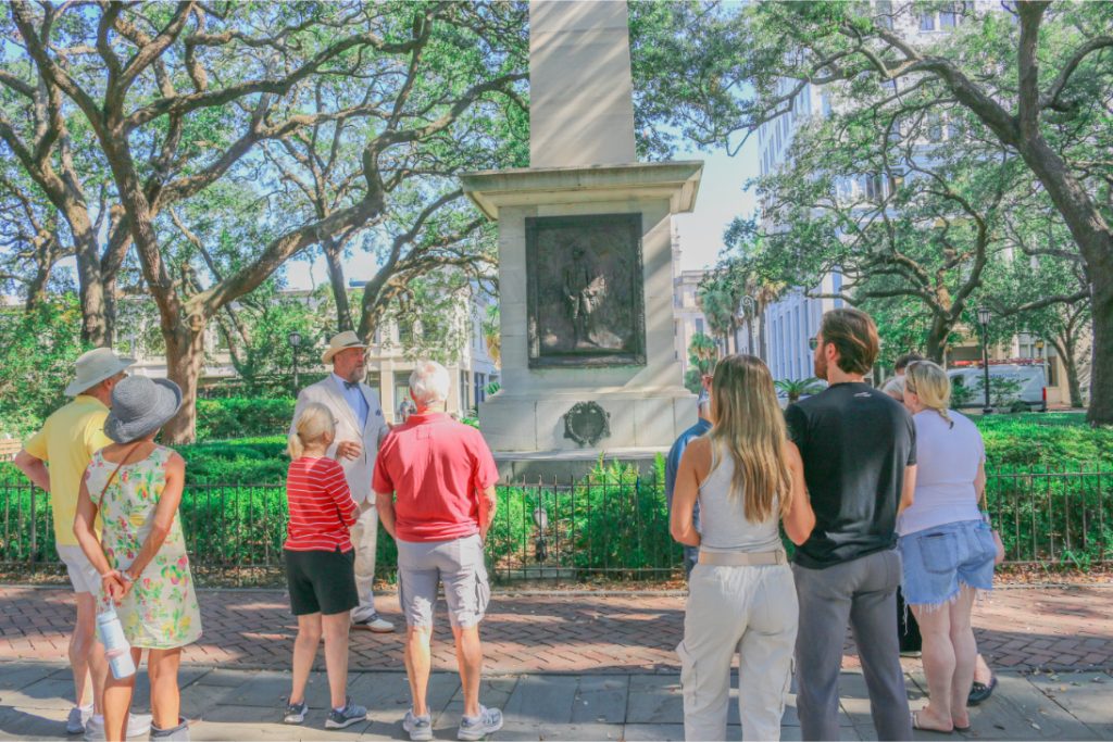 Tour group listening to a guide near a monument in a park with lush trees surrounding them