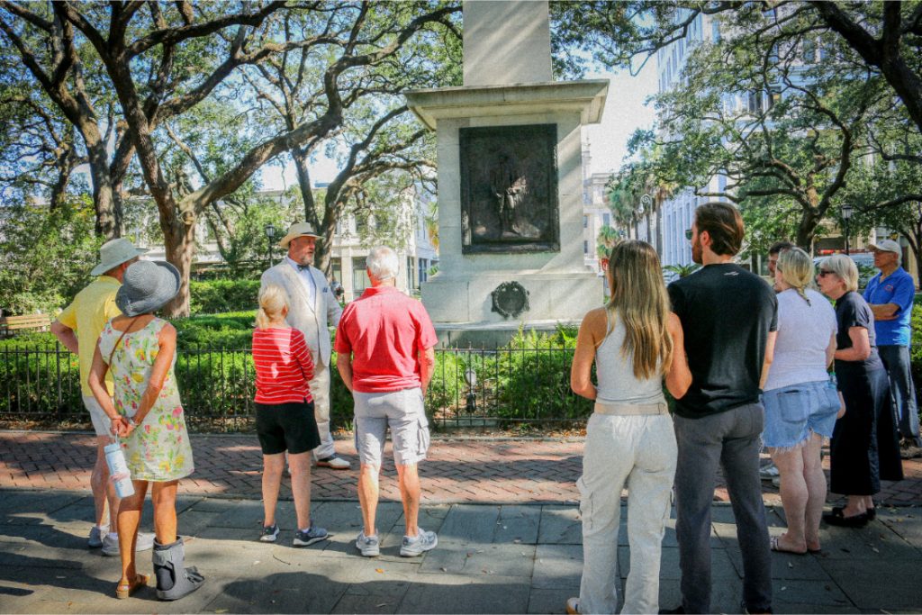 Tour group listens to guide near historic monument in a shaded park setting with trees and sunlight filtering through