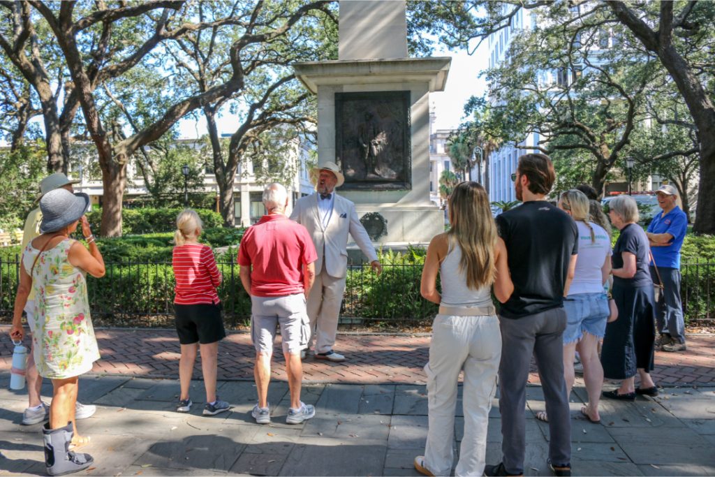 Group of people on a guided tour in a park, listening to a man near a monument on a sunny day