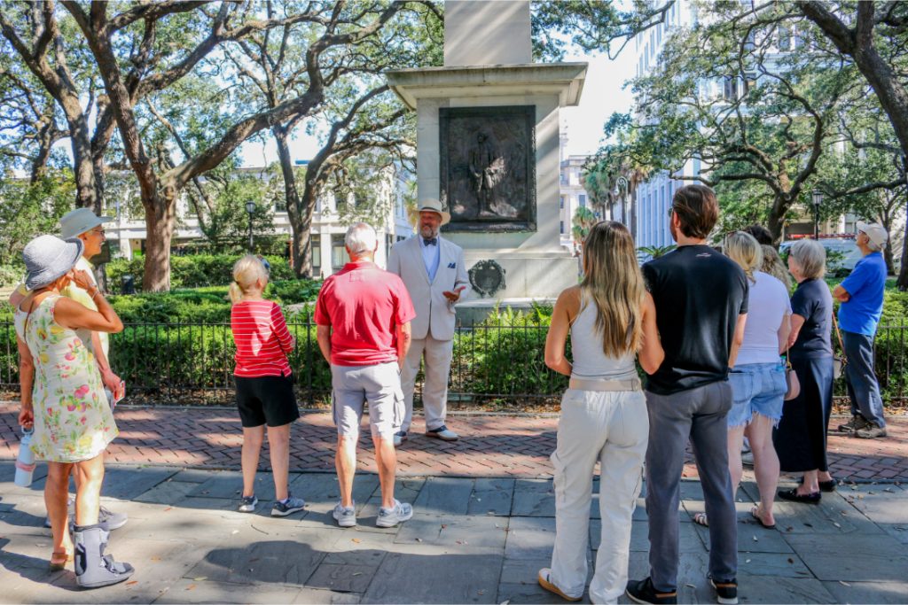 Tour group exploring historic monument in a lush park setting on a sunny day with a guide in a white suit and hat
