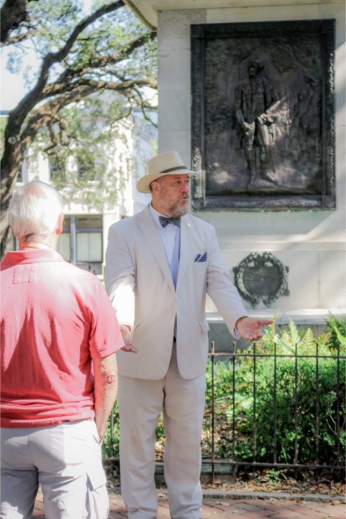 Man in a suit giving a tour in a park beside a historical monument plaque