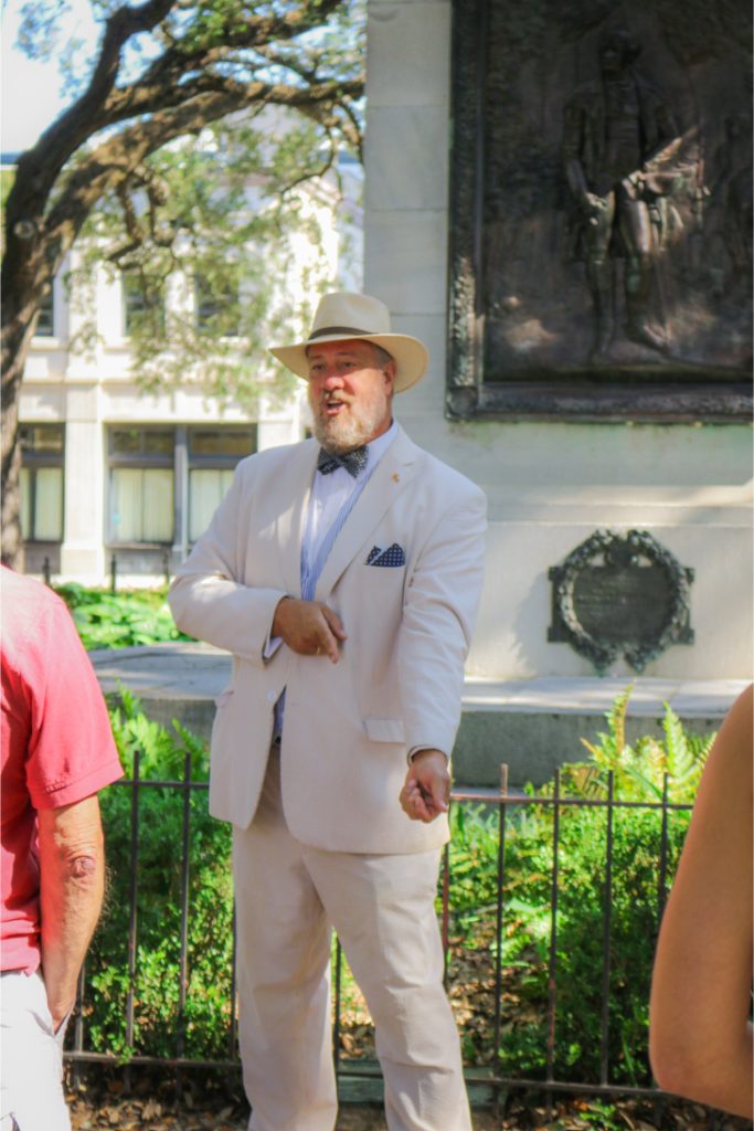 Man in a white suit and hat giving a lively tour in a garden with historic monument in the background