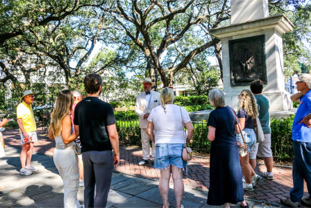 Tour group gathers around a guide in a park with a statue surrounded by tall trees in the background