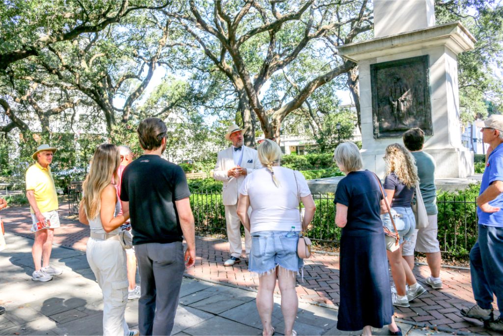 Tour guide speaking to a group of tourists in a park under large trees near a historic monument