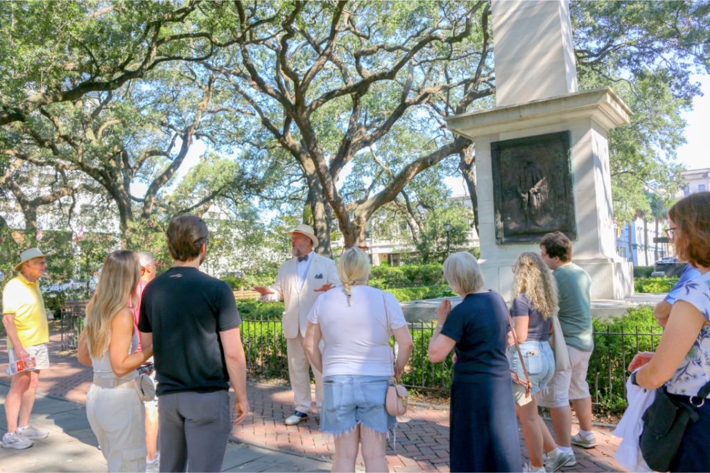 Tour group listens to a guide wearing a hat in a leafy park setting with a statue nearby