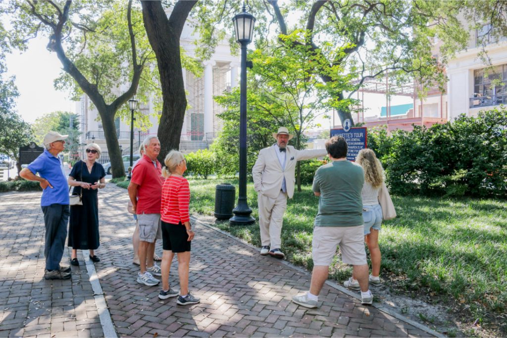Tour guide in white suit leads group on historical walking tour in a park with lush greenery and historic buildings