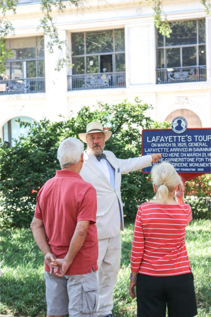 Tour guide in beige suit explaining Lafayette's 1825 Savannah visit to senior tourists in a garden setting