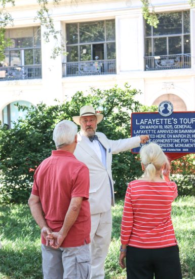 Tour guide in white suit explaining historic Lafayette's Tour sign to two older adults in a garden setting