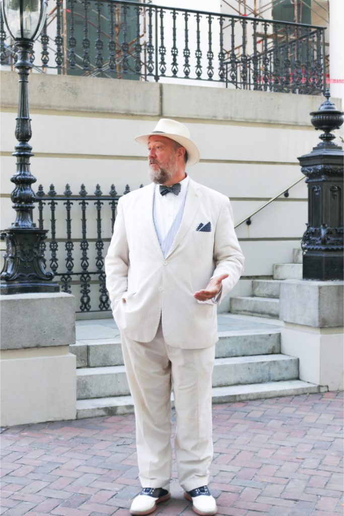 Man in white suit and hat standing on cobblestone street near vintage lamp posts and iron railings