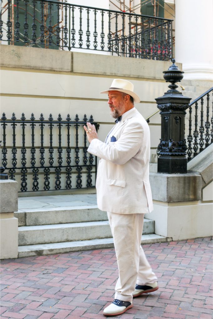 Man in white suit and hat walking on brick path, urban background with iron railings and steps