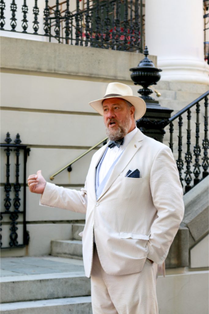 Man in a white suit and hat gesturing on a staircase outside an elegant building