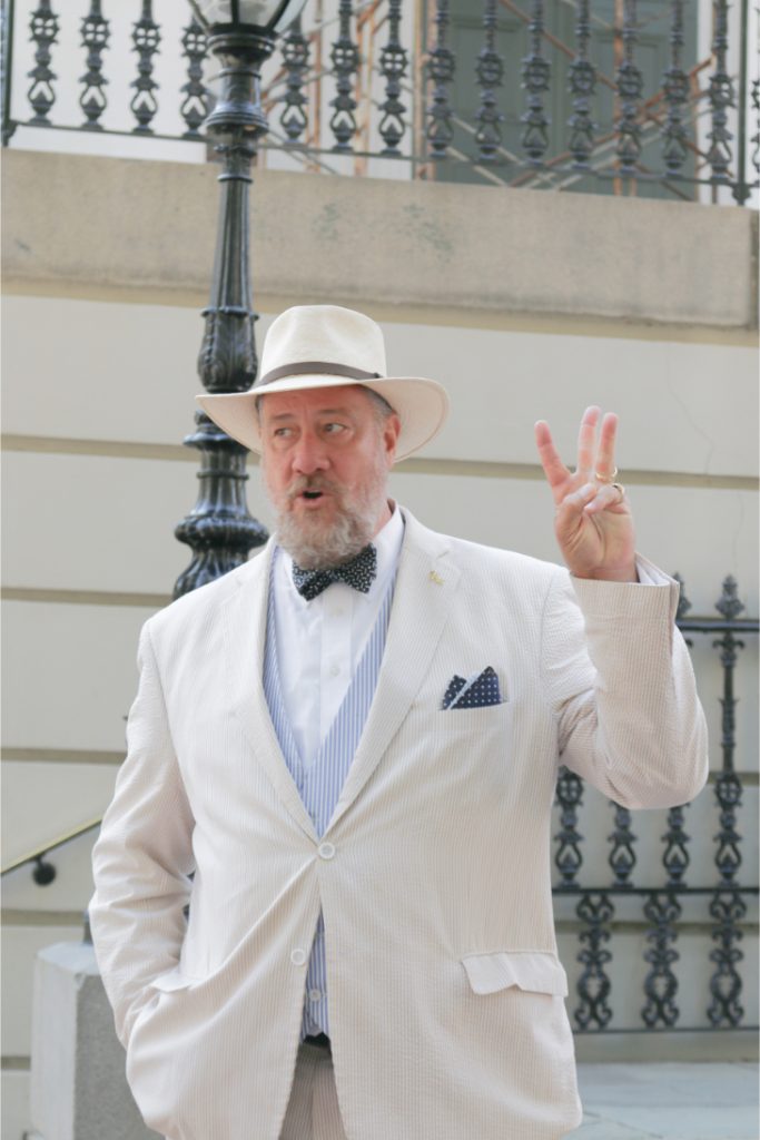 Man in white suit and hat gesturing with two fingers, standing near a decorative railing outdoors