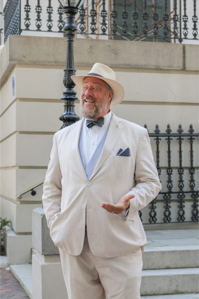 Man in a stylish suit and hat stands outdoors, gesturing with a smile, near a decorative railing