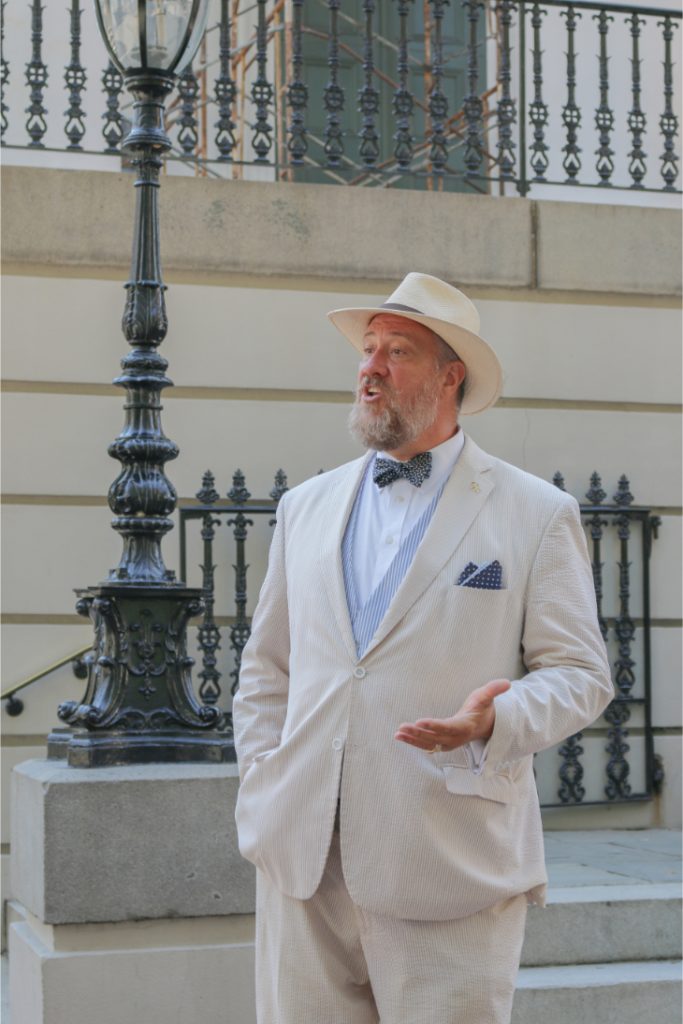 Man in white suit and hat giving a speech outdoors near ornate railing and lamp post