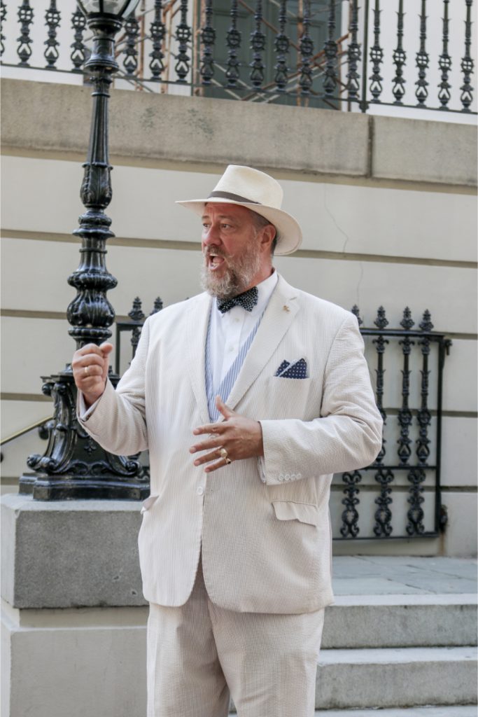 Man in stylish white suit and hat gestures passionately outside a historic building steps lamp post in background