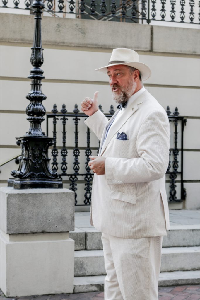 Man in white suit and hat gestures confidently while standing near a decorative lamppost and wrought iron fence