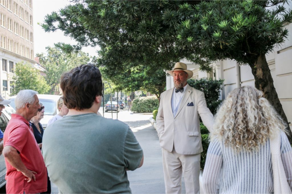 Group gathered outdoors, listening to a man in a white suit and hat, with trees and buildings in the background