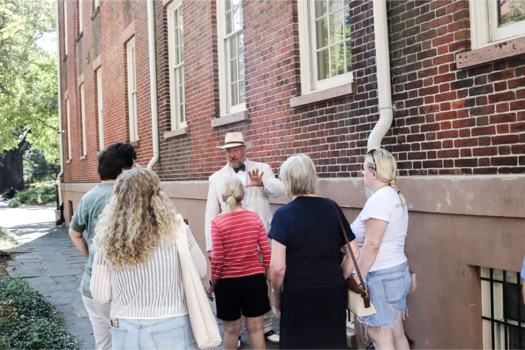 Group on historical walking tour listens to guide in white hat outside brick building