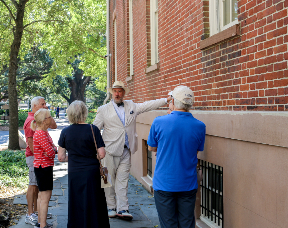 Tour guide in a suit and hat leading a group near a historic brick building under trees