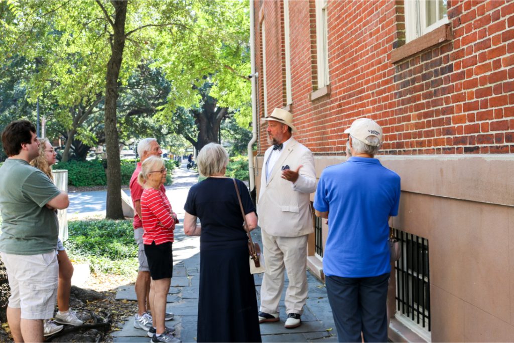 Tour guide in white suit leads group on historic brick wall tour, surrounded by trees and lush greenery