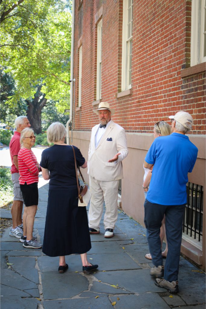 Man in white suit with hat leading a guided outdoor tour for a diverse group by a historic brick building
