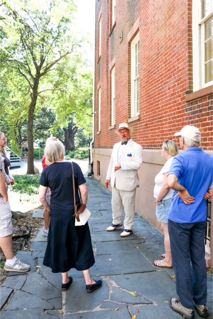 Tour guide in fedora leading a group by a historic brick building on a sunny day