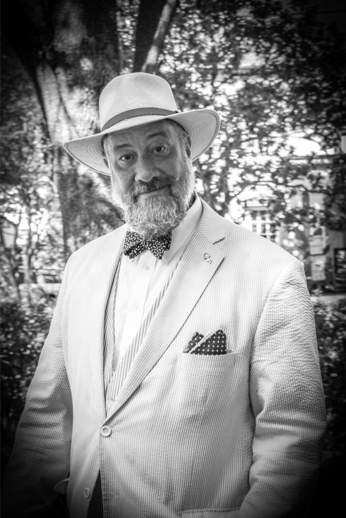 Man in a suit and hat with a beard and bow tie smiles outdoors in black and white photograph