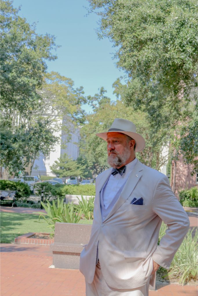 Man in a light suit and hat stands in a park during daytime with trees and buildings in the background