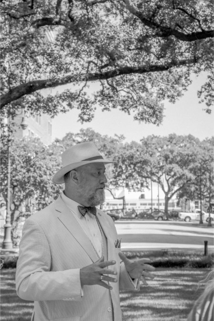 Man in a suit and hat speaks outdoors, surrounded by trees, in black and white photography ambiance