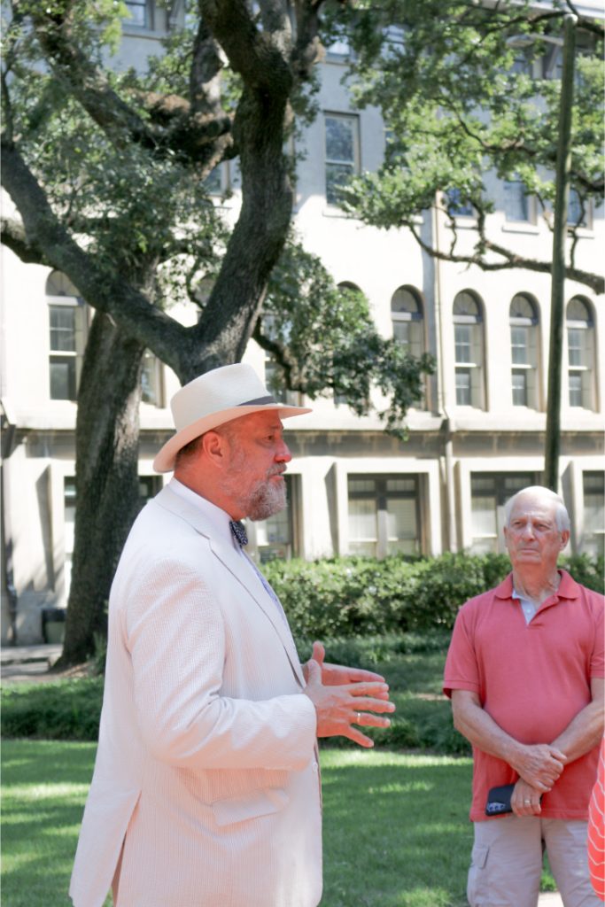 Man in white suit and hat speaking outdoors in a park setting with trees and historic building in background