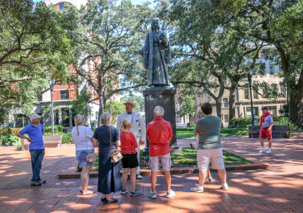 Tour group listening to a guide near a historic statue in a sunlit park with trees and buildings in the background