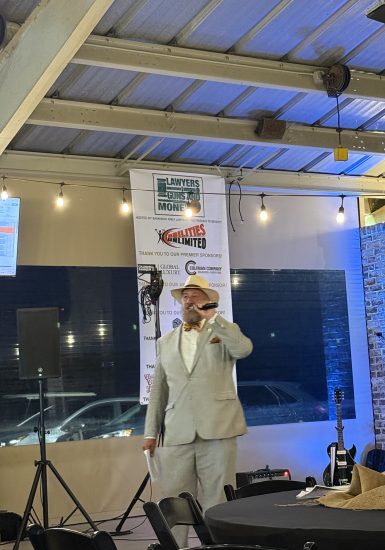 Savannah Dan in a suit and hat speaks at an indoor event with a banner for Lawyers Guns and Money in the background