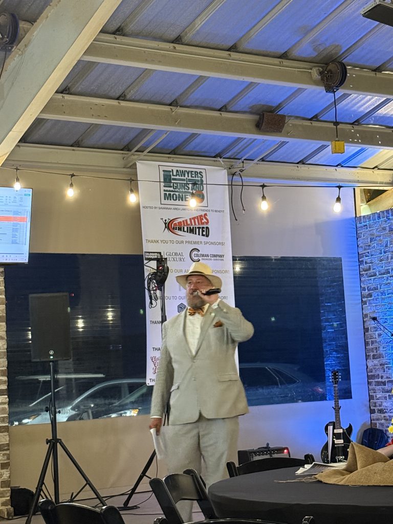 Savannah Dan in a suit and hat speaks at an indoor event with a banner for Lawyers Guns and Money in the background