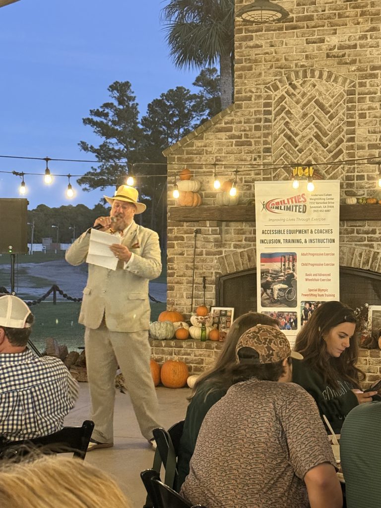 Man in suit with hat speaking at outdoor event by fireplace with pumpkins, banner about accessibility and inclusion programs