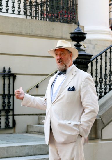 Man in a light suit and hat talking on historic stone steps with ornate railing background